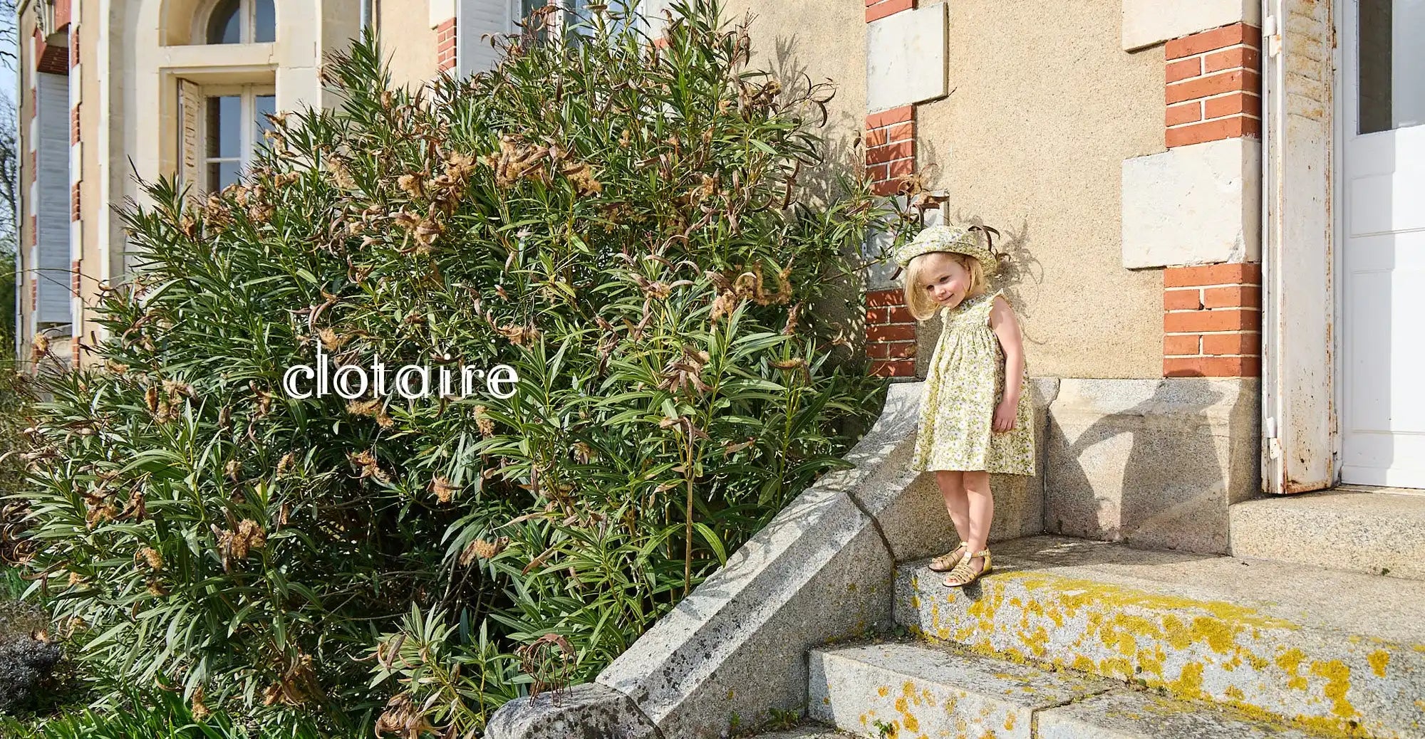 Un enfant portant une robe et un chapeau se tient sur des marches en béton près d'un buisson de lauriers-roses.